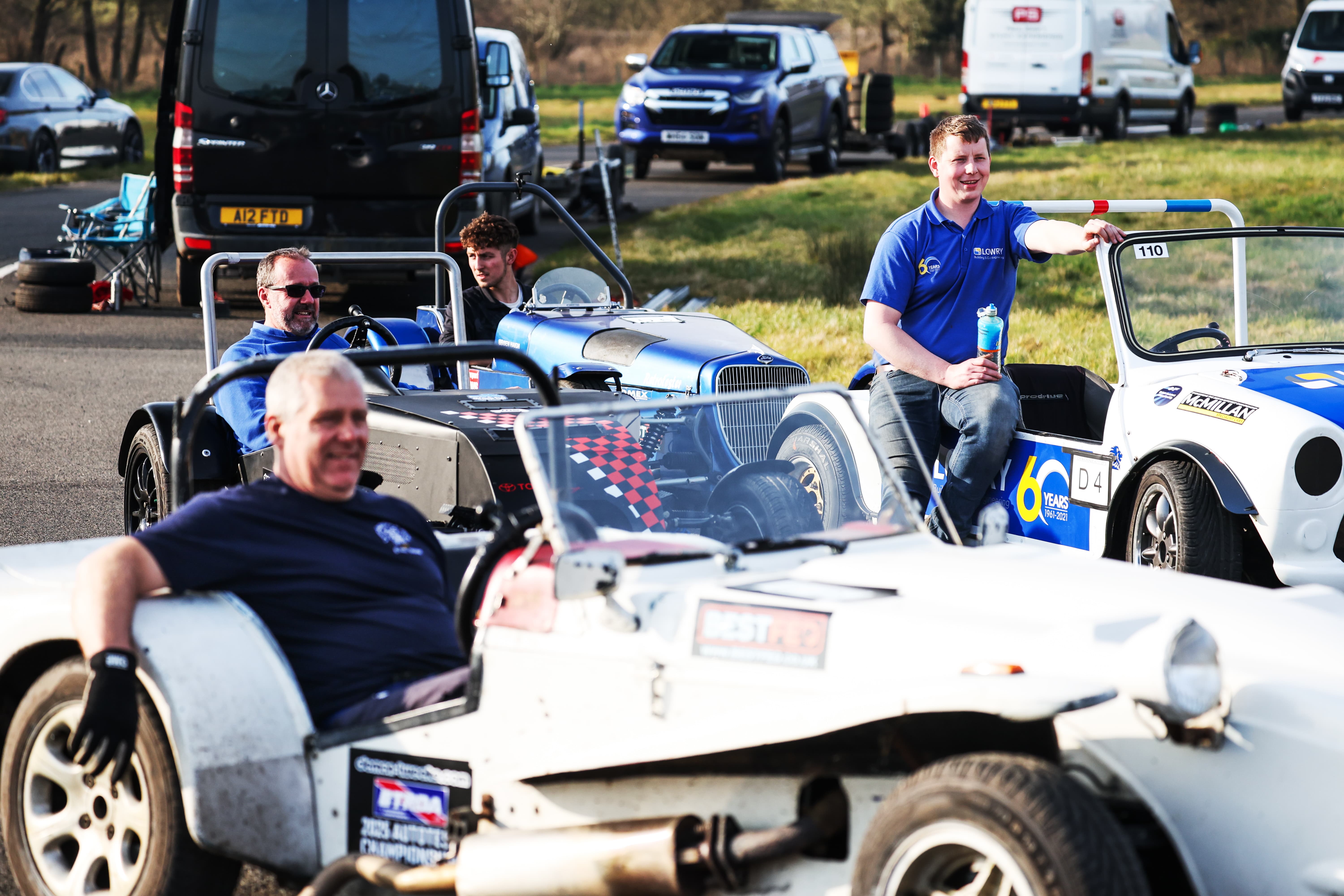 Four men sitting in three different vintage-style open-top race cars in an outdoor parking area, with vehicles and green grass in the background.