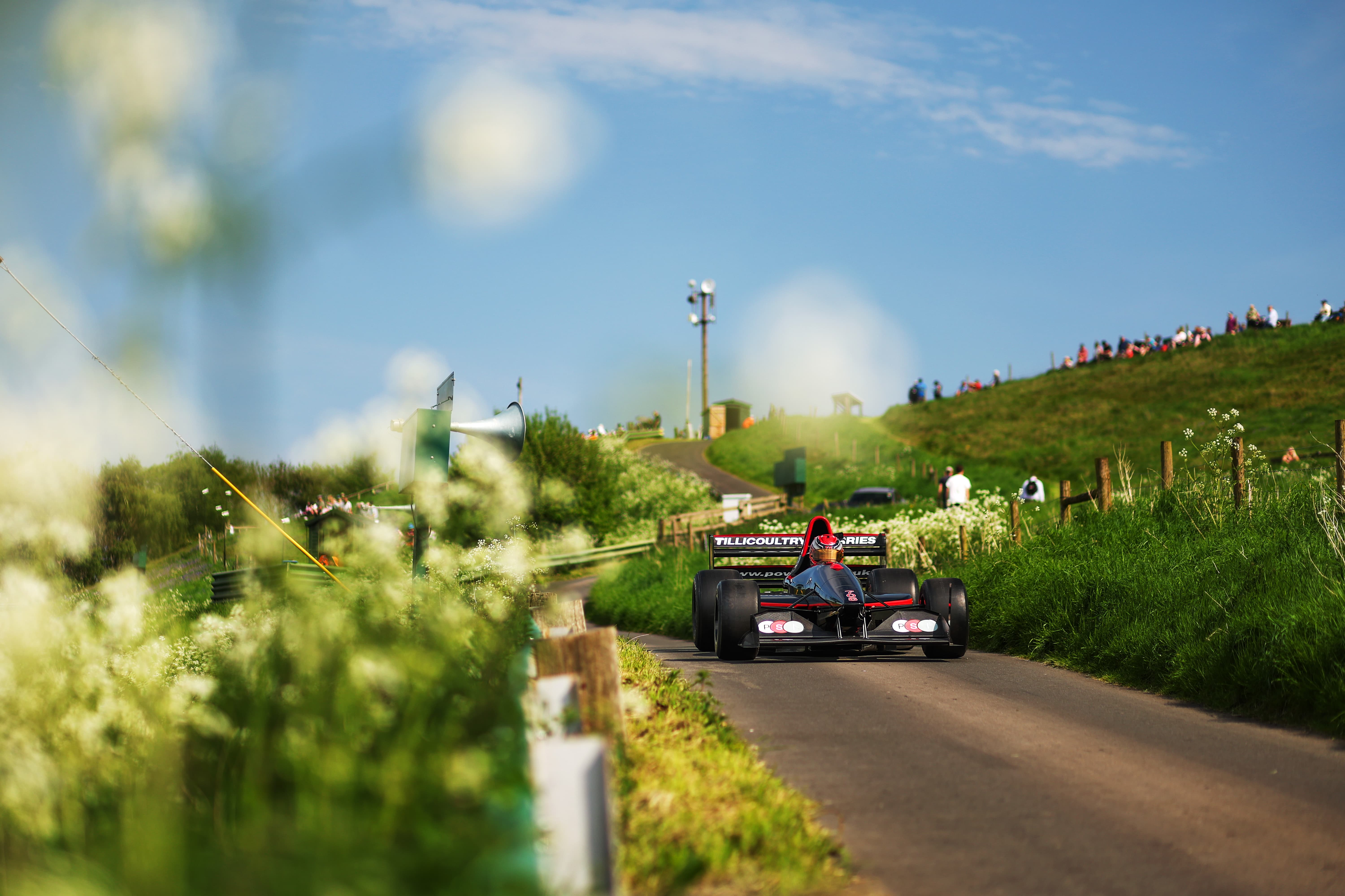 Racecar driving on a narrow country road with green hills and spectators in the background.