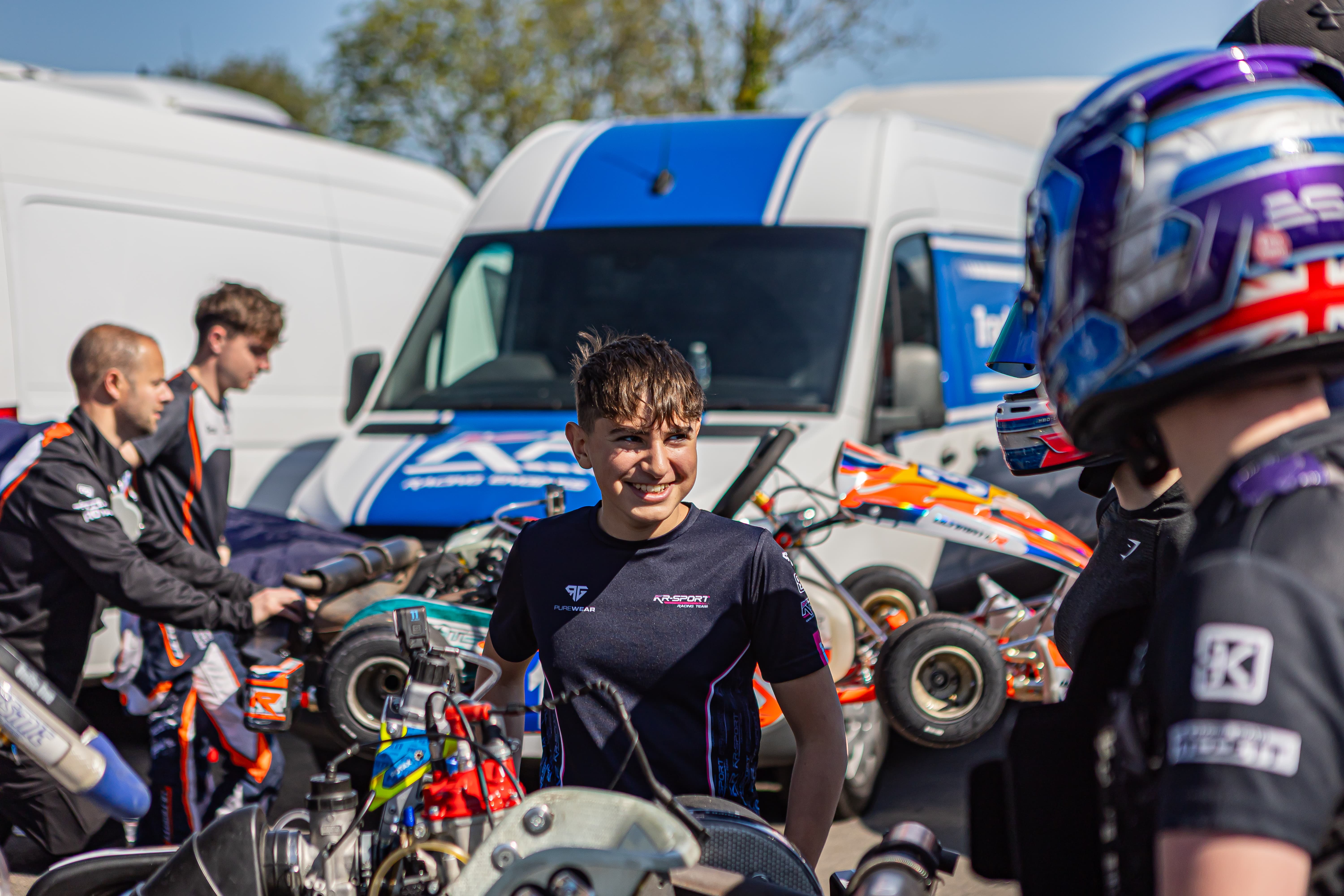 Young man smiling among go-kart racers and mechanics working near equipment outdoors with vans in background.