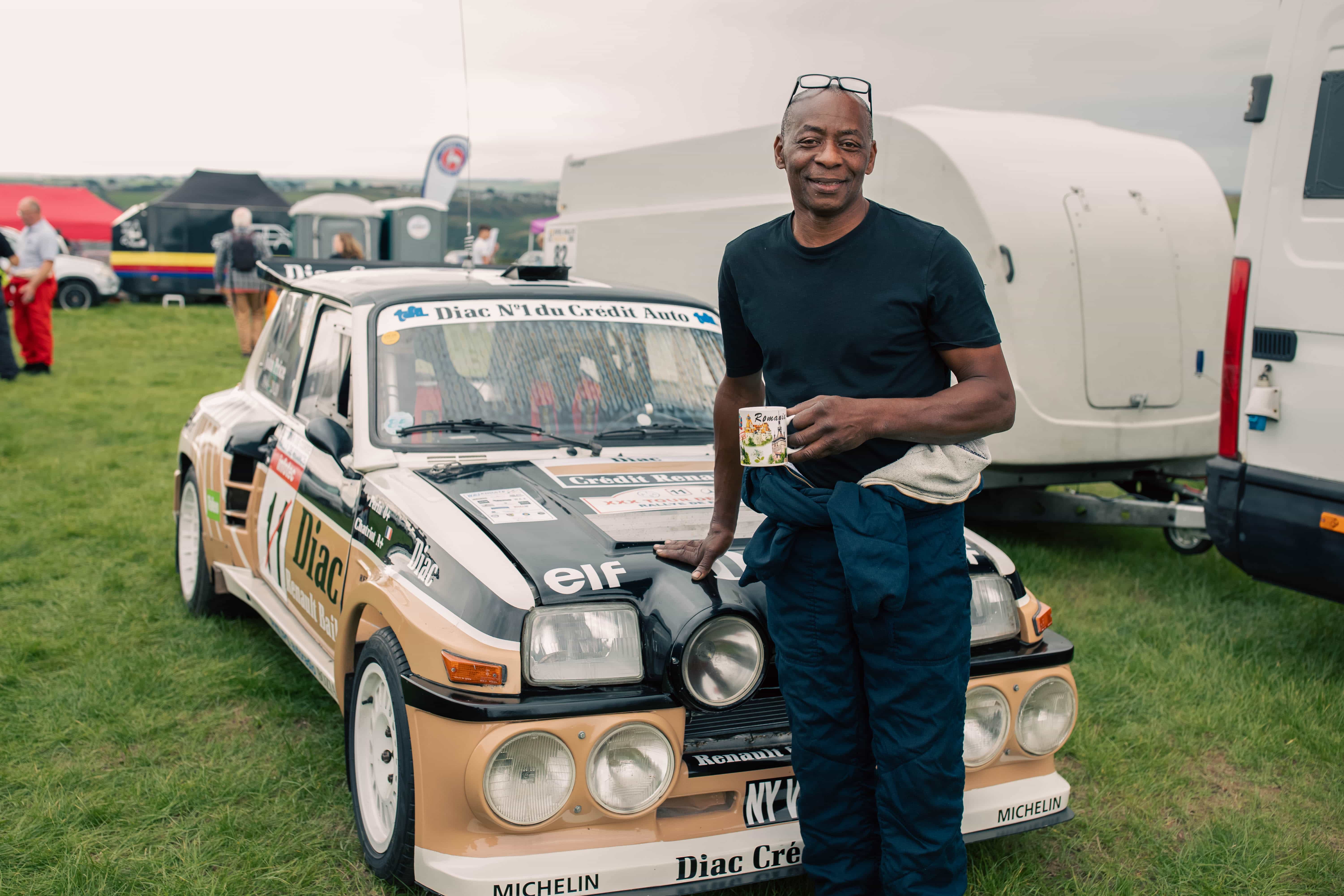 Man in blue racing suit holding a decorated mug, leaning against a vintage race car on a grassy field.