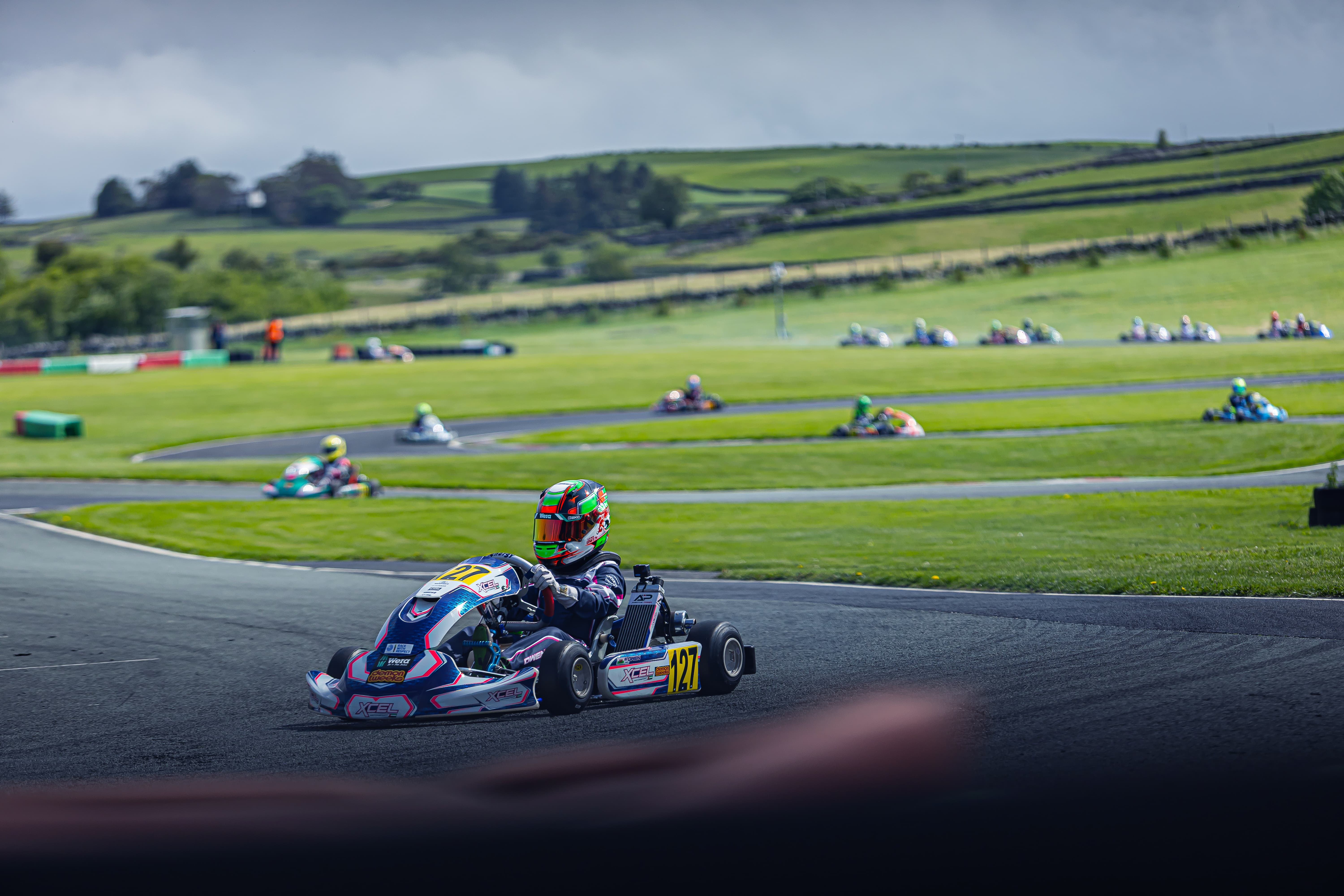 Go-kart racer in blue and red kart number 127 navigating a turn on a racetrack with other racers and green hills in the background.