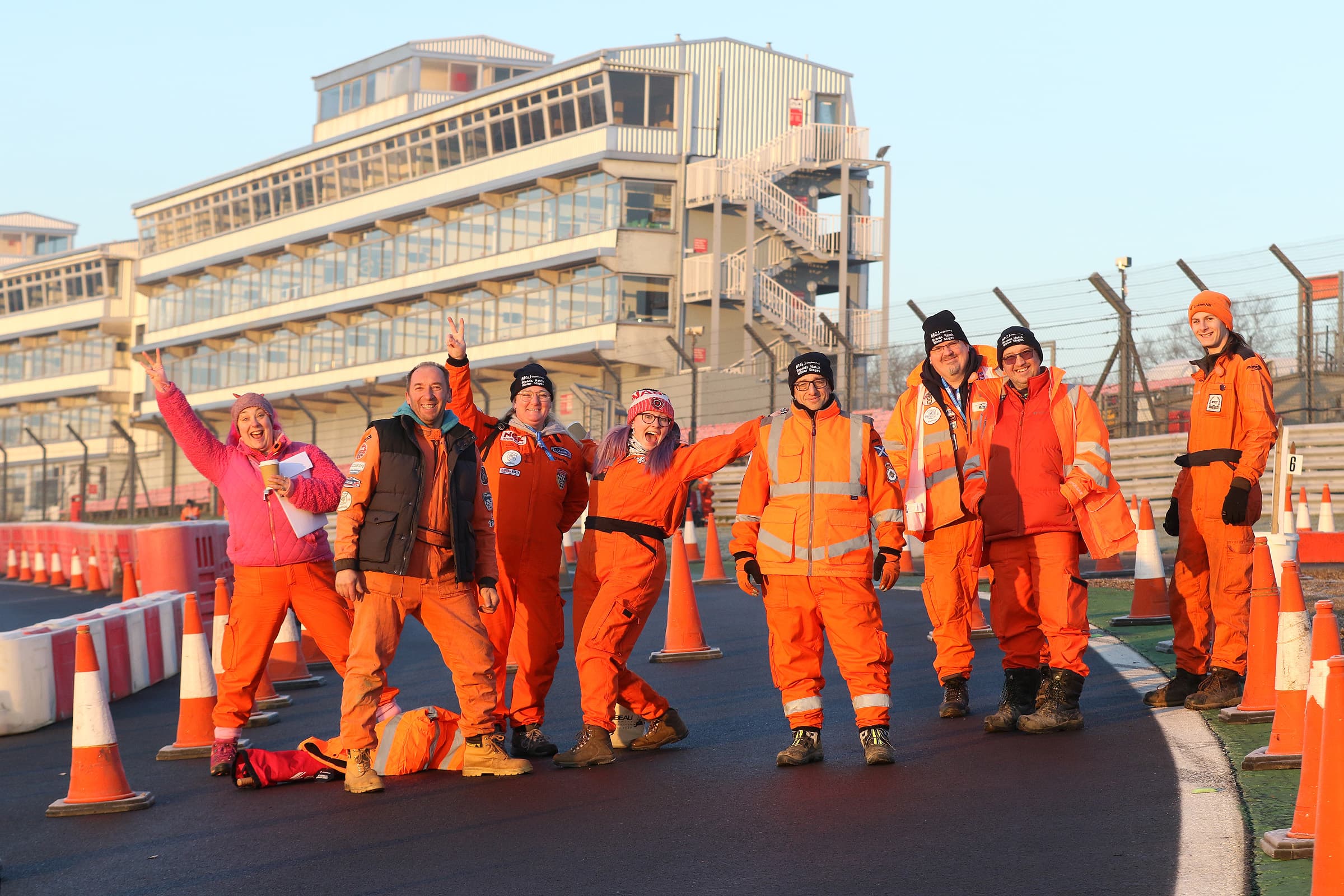 Group of nine race marshals in orange safety uniforms standing on a racetrack with a multi-story building in the background.
