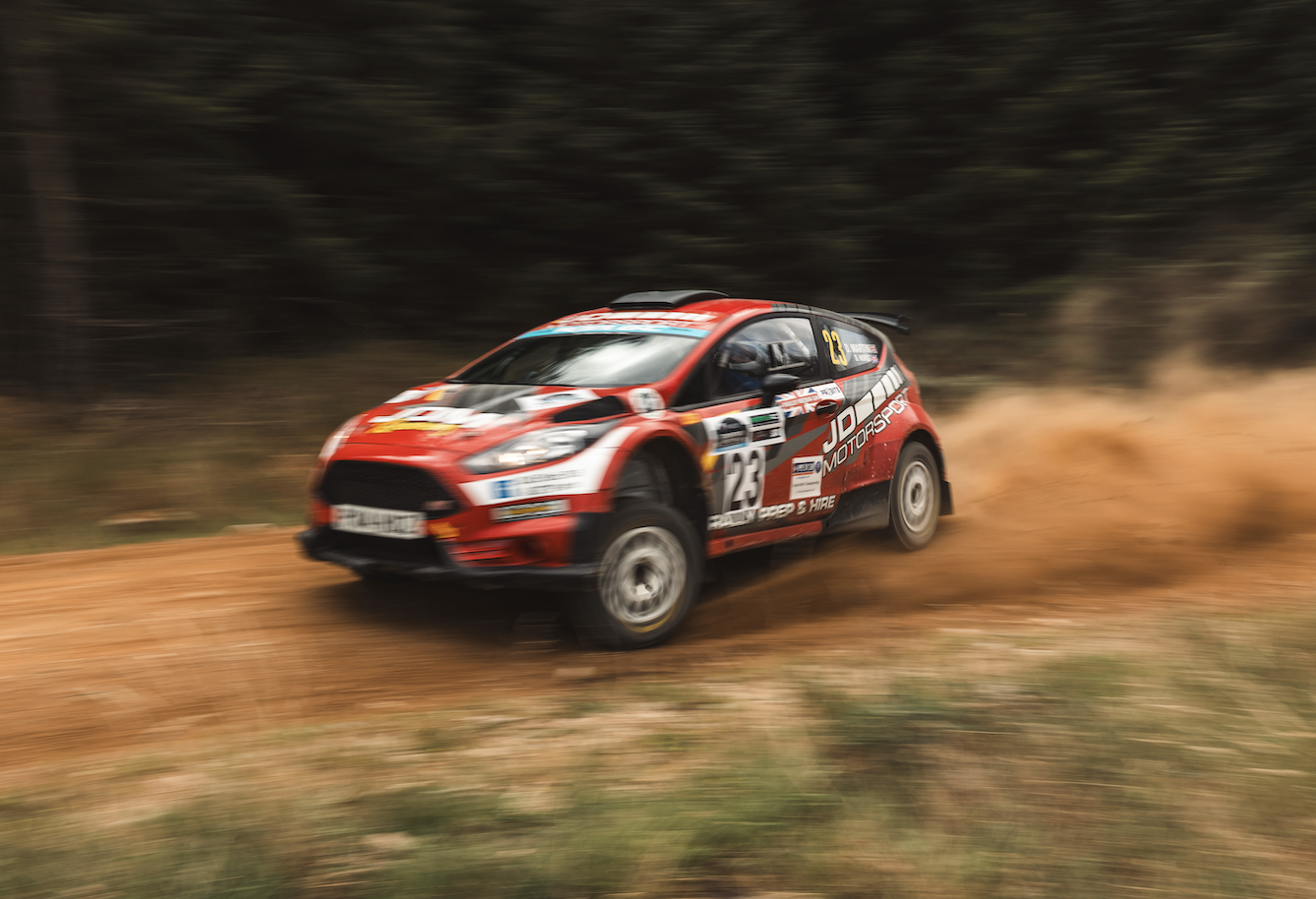 Red rally car number 23 speeding on a dirt road, kicking up a cloud of dust with a blurred forest background.
