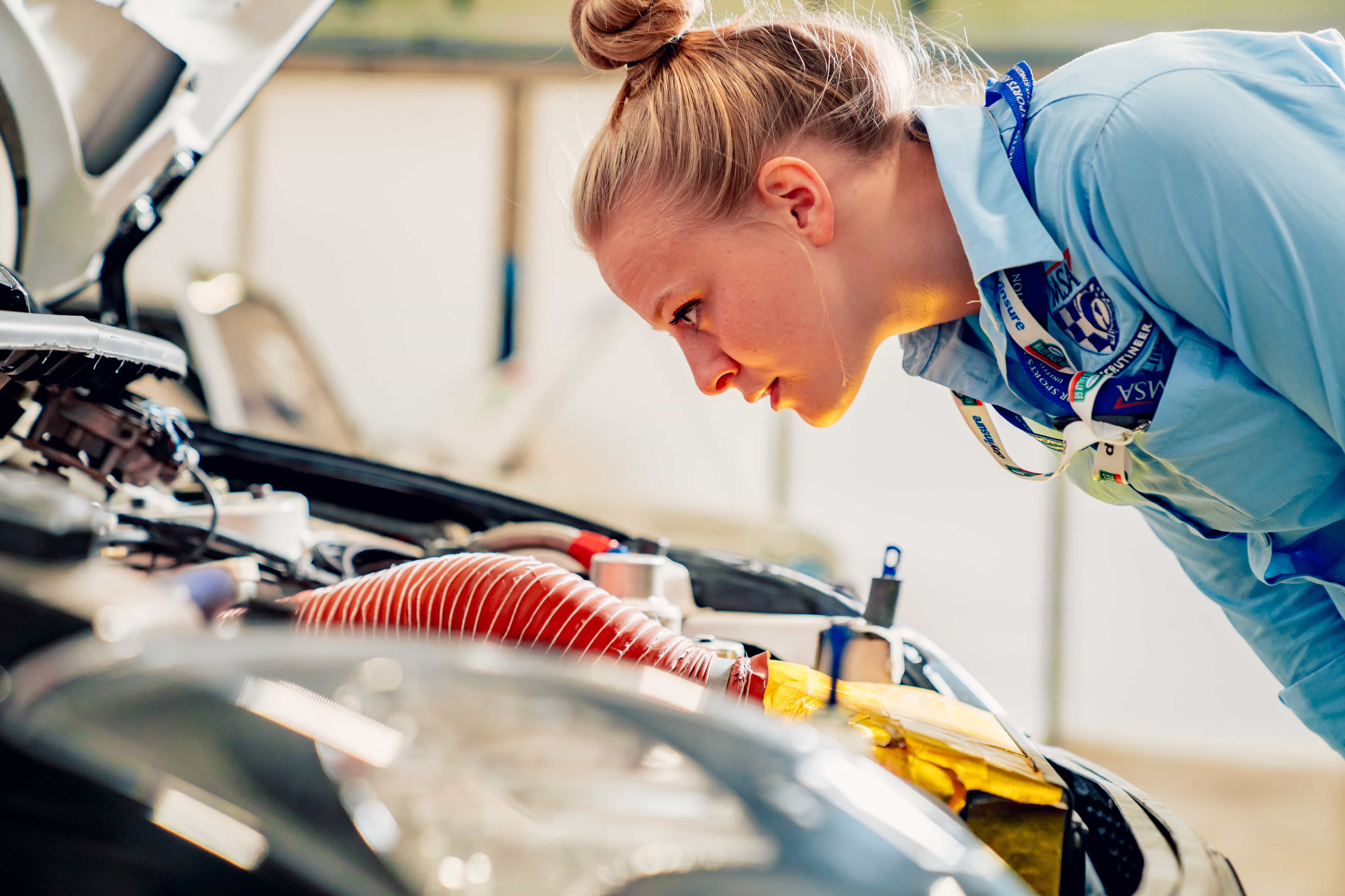 Woman wearing a blue shirt and lanyard closely inspecting an open car engine under the hood.
